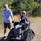 Volunteer walking alongside the track chair on a dirt trail with participant waving