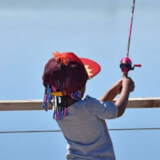 A little girl holds a fishing pole over a railing wearing a red hat.