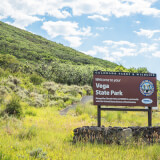 The Vega State Park road sign is viewed against a prairie hilltop background with cloudy blue sky.